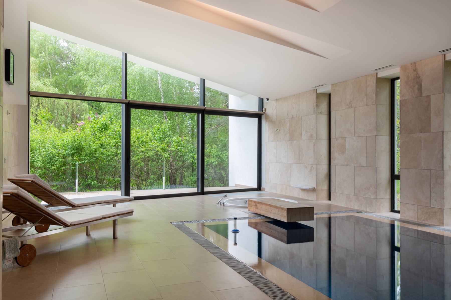 Espace piscine intérieur équipé : bassin encadré de travertin, éclairage et grandes baies vitrées sur la végétation